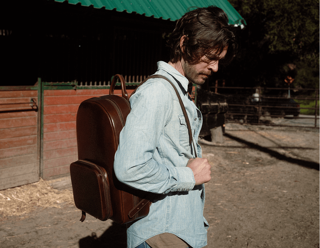 Man wearing a brown leather backpack walking past a stable