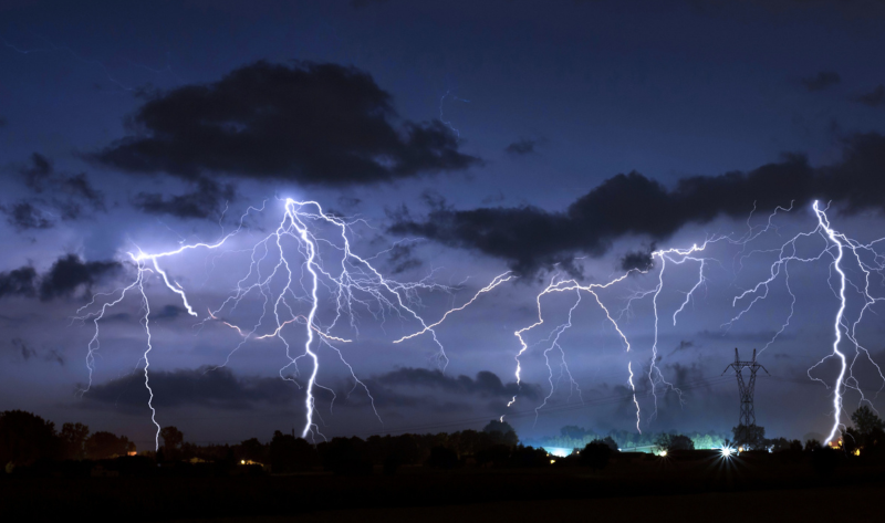 Multiple lightning bolts strike across a dark sky above a landscape with trees, buildings, and an electrical tower at night.