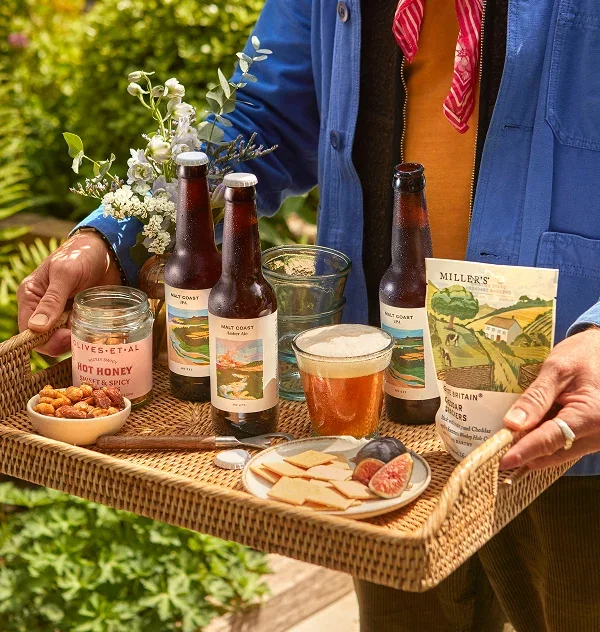 A close-up of a person carrying a woven rattan tray loaded with an artisan beer and snack hamper in a sunlit garden. The tray holds three bottles of ‘Malt Coast’ craft beer, a poured glass of amber ale, a packet of ‘Miller’s Cheddar Crackers’, a jar of ‘Olives ET AL Hot Honey Nuts’, and a small plate of crackers with fresh sliced figs.