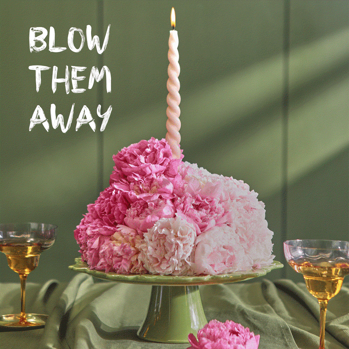 A creative birthday display featuring a mound of fluffy pink and pale cream peonies arranged on a green ceramic cake stand to resemble a celebration cake. A single, tall pink twisted candle stands lit at the top that then gets blown out. Two glasses of champagne flank the stand on a draped sage green tablecloth, with text in the top left corner reading ‘BLOW THEM AWAY’.