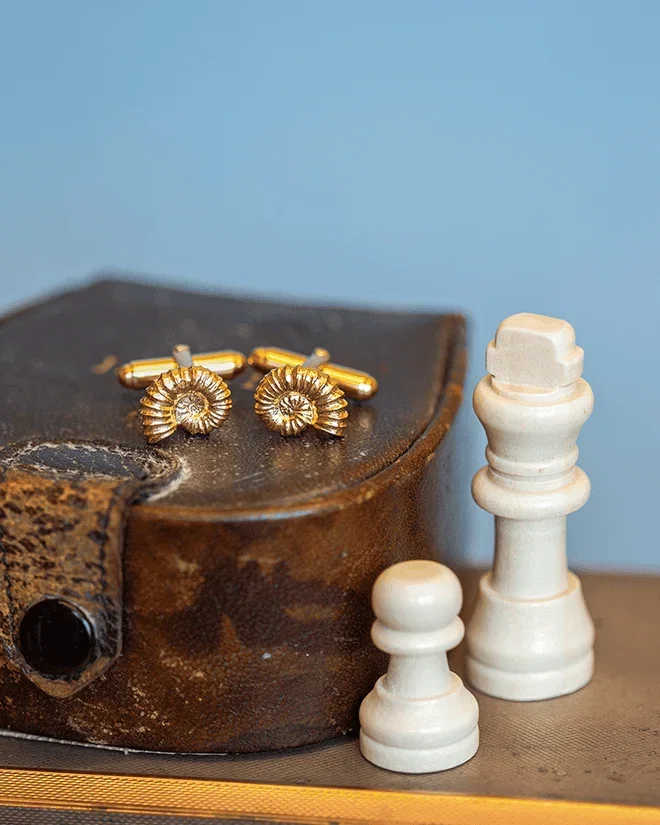 Alex Monroe Gold ammonite cufflinks displayed beside chess pieces on a vintage chessboard.