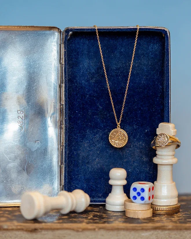 Alex Monroe Gold Nautical Antique Coin Necklace and Signet Ring in a vintage presentation box beside chess pieces and a dice