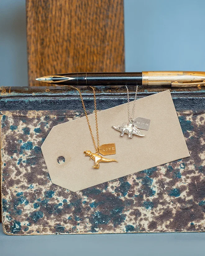 Alex Monroe Gold Tyrannosaurus Rex Necklace and Silver Stegosaurus Necklace resting on a vintage metal box with a fountain pen above