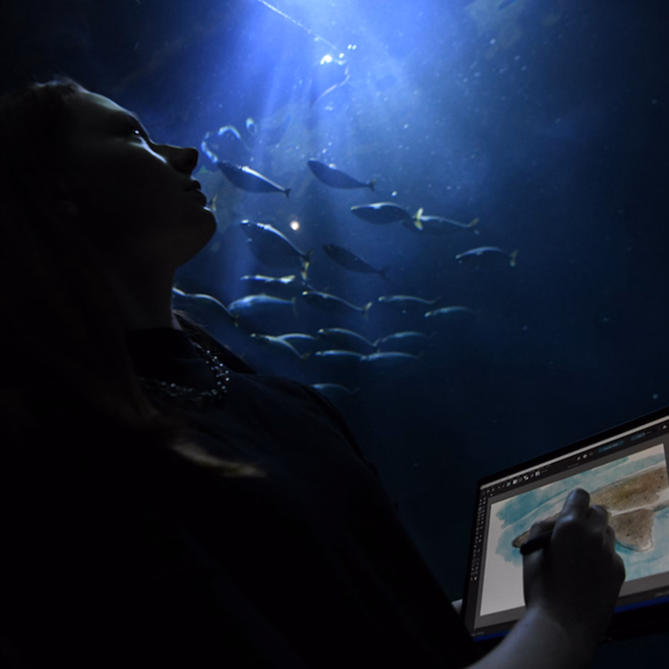 Natalie Donato sketching on a laptop while seated beside a glowing aquarium, with fish swimming in blue light overhead.