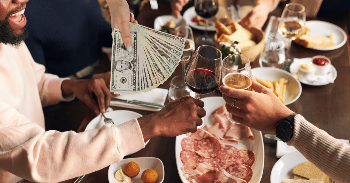 Two customers at restaurant clink wine glasses over an array of food while a hand brandishes several paper bills.