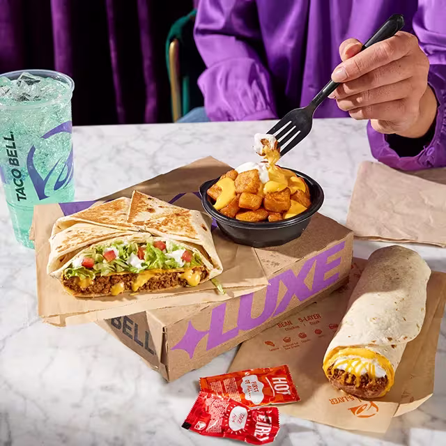 An image of someone holding a fork and enjoying various Taco Bell menu items laid across a table.
