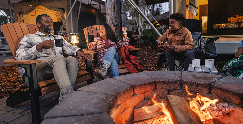 Three people sitting around a campfire enjoying drinks.