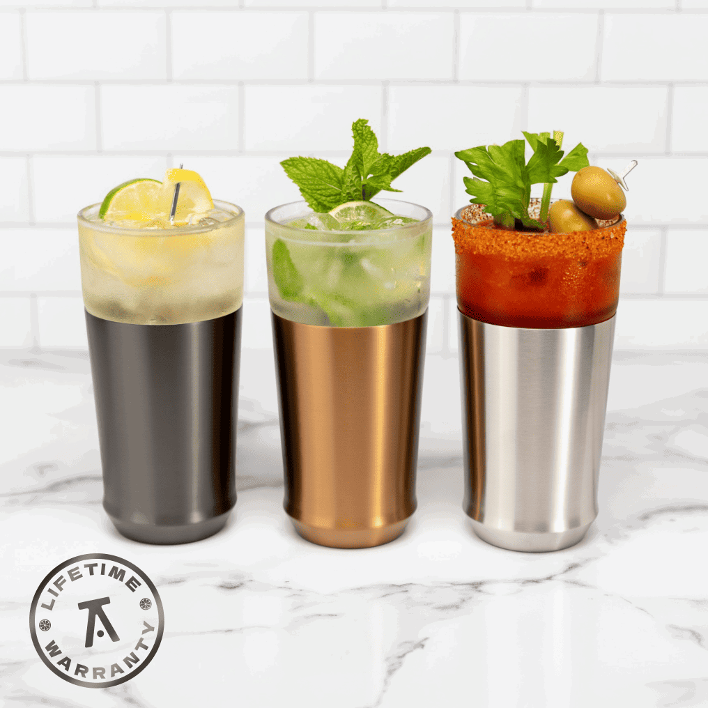 Three Elevated Craft Hybrid Pint Glasses in gunmetal, copper, and stainless steel finishes, each filled with a different cocktail: a citrus drink, a mojito, and a Bloody Mary with a Lifetime Warranty badge