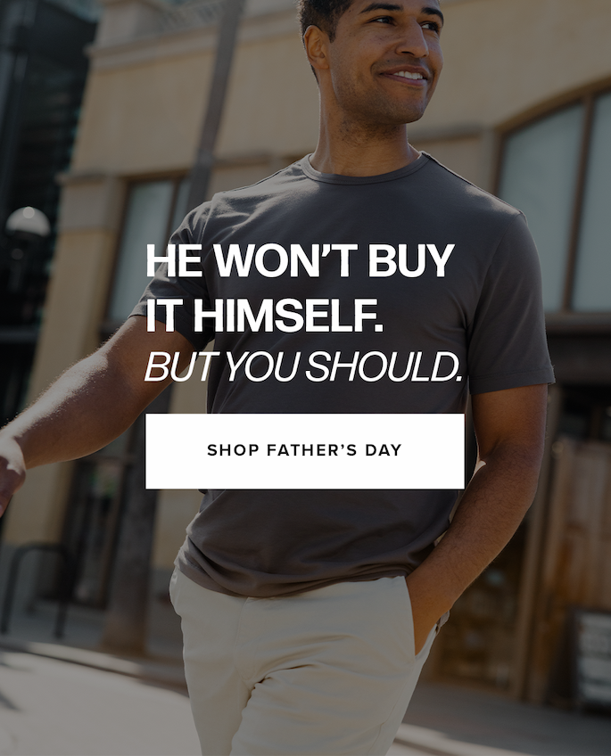 A man walks outdoors in a dark grey t-shirt and tan pants with a blurred city street background. Text reads: 'He won’t buy it himself. But you should. Shop Father’s Day'