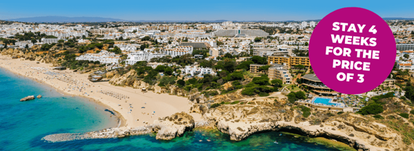 Aerial view of a sandy beach with clear blue water and coastal buildings in the background.