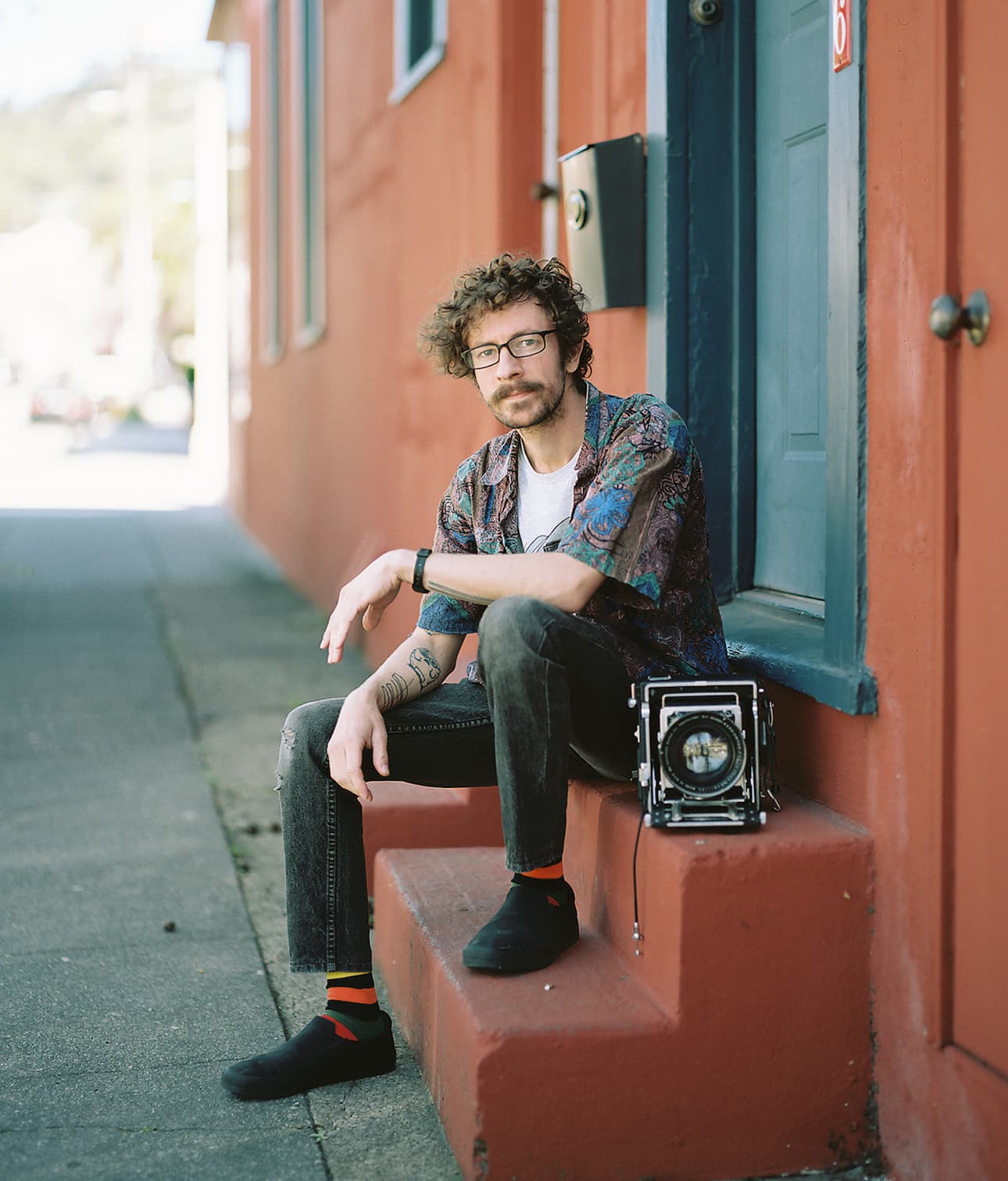 Dave Rollans sits on a doorstep with his Graflex 4x5 Speed Graphic large format camera by his side.