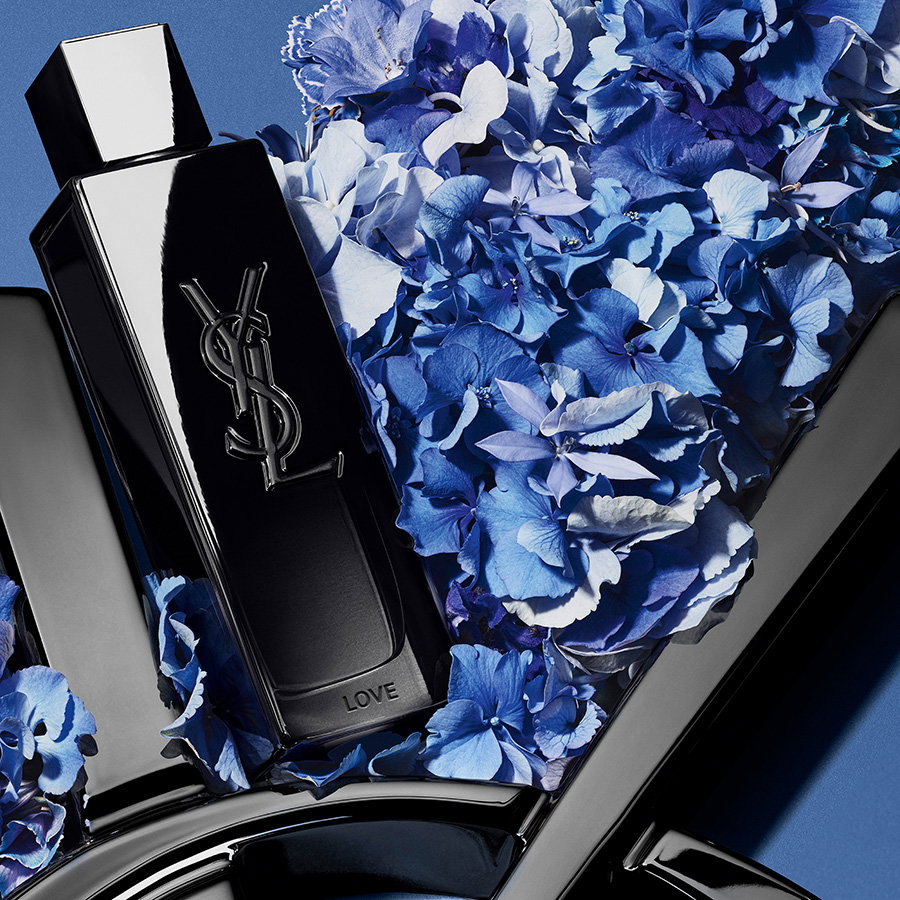 A sleek black YSL fragrance bottle engraved with the word "LOVE", nestled in a bed of vibrant blue hydrangeas.