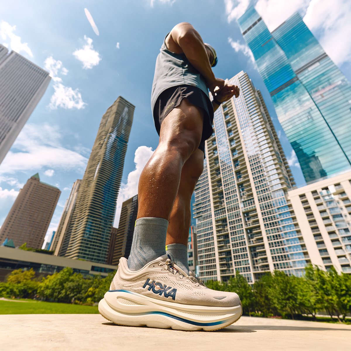 Low-angle image of a man wearing HOKA shoes. In the background, there are city buildings and a blue sky.