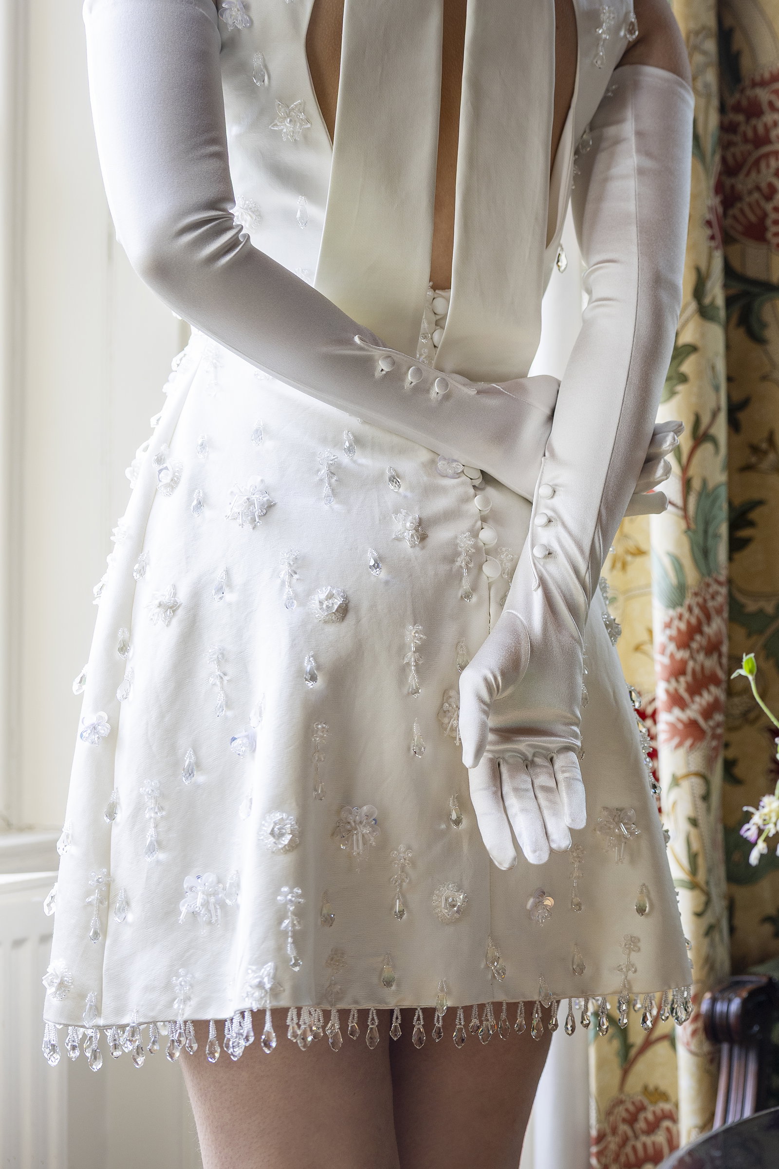 A close up of woman crossing her arms behind the back wearing long satin gloves in ivory standing by the window