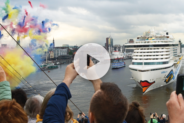 Menschen stehen am Hafen und fotografieren ein großes Kreuzfahrtschiff, das an einem Fluss vorbeifährt; links steigen bunte Rauchwolken auf, im Hintergrund sind Gebäude unter bewölktem Himmel sichtbar.