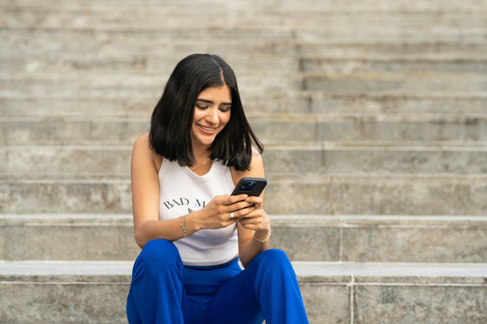 A smiling woman sitting on outdoor stone steps, looking at her smartphone.