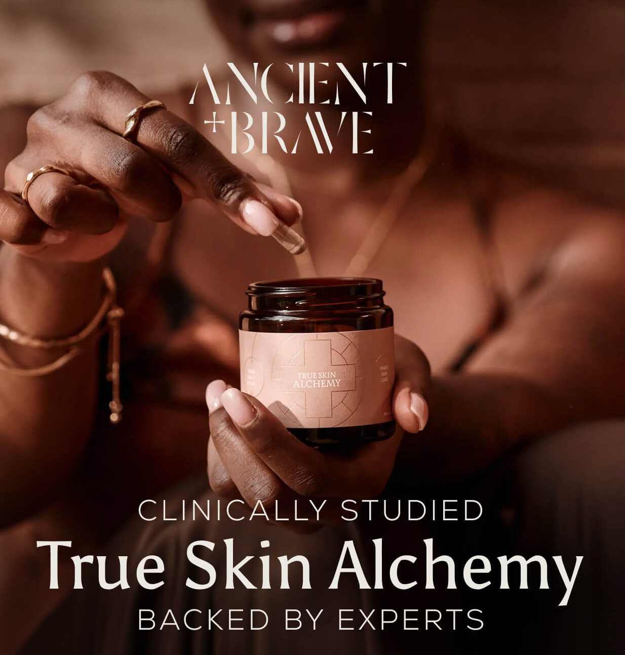 An Ancient + Brave campaign image. A woman holds an open jar of True Skin Alchemy, with a dropper held above it in her other hand. The text reads: "Clinically Studied — True Skin Alchemy — Backed by Experts."