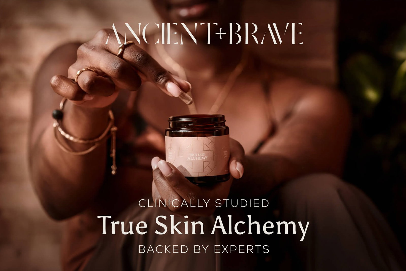 An Ancient + Brave campaign image. A woman holds an open jar of True Skin Alchemy, with a dropper held above it in her other hand.  The text reads: "Clinically Studied — True Skin Alchemy — Backed by Experts."