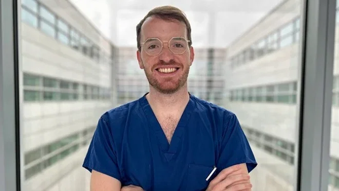 Picture of Dr Michael Wilkinson, a new Action Medical Research Research Training Fellow. He is wearing navy blue clinical uniform, and is standing with his arms folded, smiling, in front of a hospital window. Other parts of a hospital building can be seen in the background.