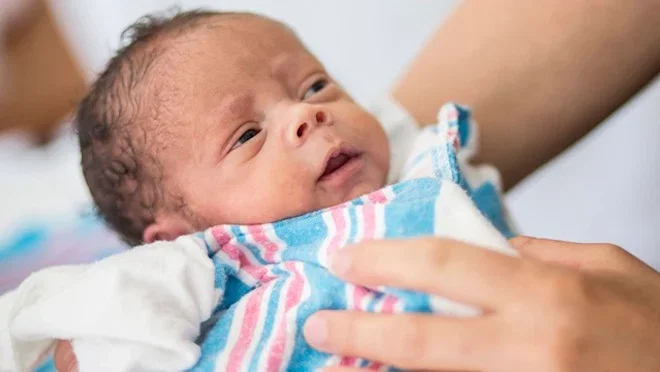 Picture of a newborn baby, with black hair, in the hands of a caregiver in a hospital setting. The baby is wrapped in a blue, pink and white stripe sheet or blanket.