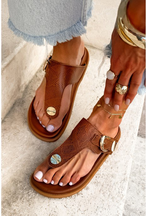 Close-up of brown textured leather thong sandals with gold buckle and coin accents on a pedicured foot, frayed-hem jeans and anklet