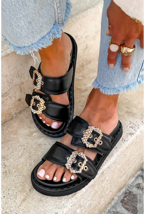 Black leather double-strap sandals with decorative gold crystal buckles on model's pedicured feet, frayed-hem jeans and rings
