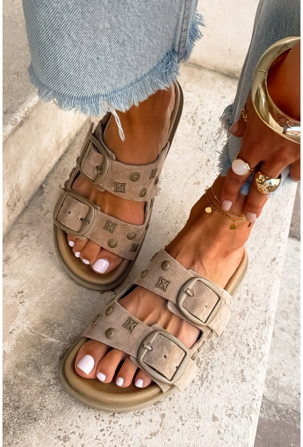 Close-up of woman's tan suede double-buckle sandals with white pedicure, gold anklet and jewellery, frayed-hem jeans on stone step