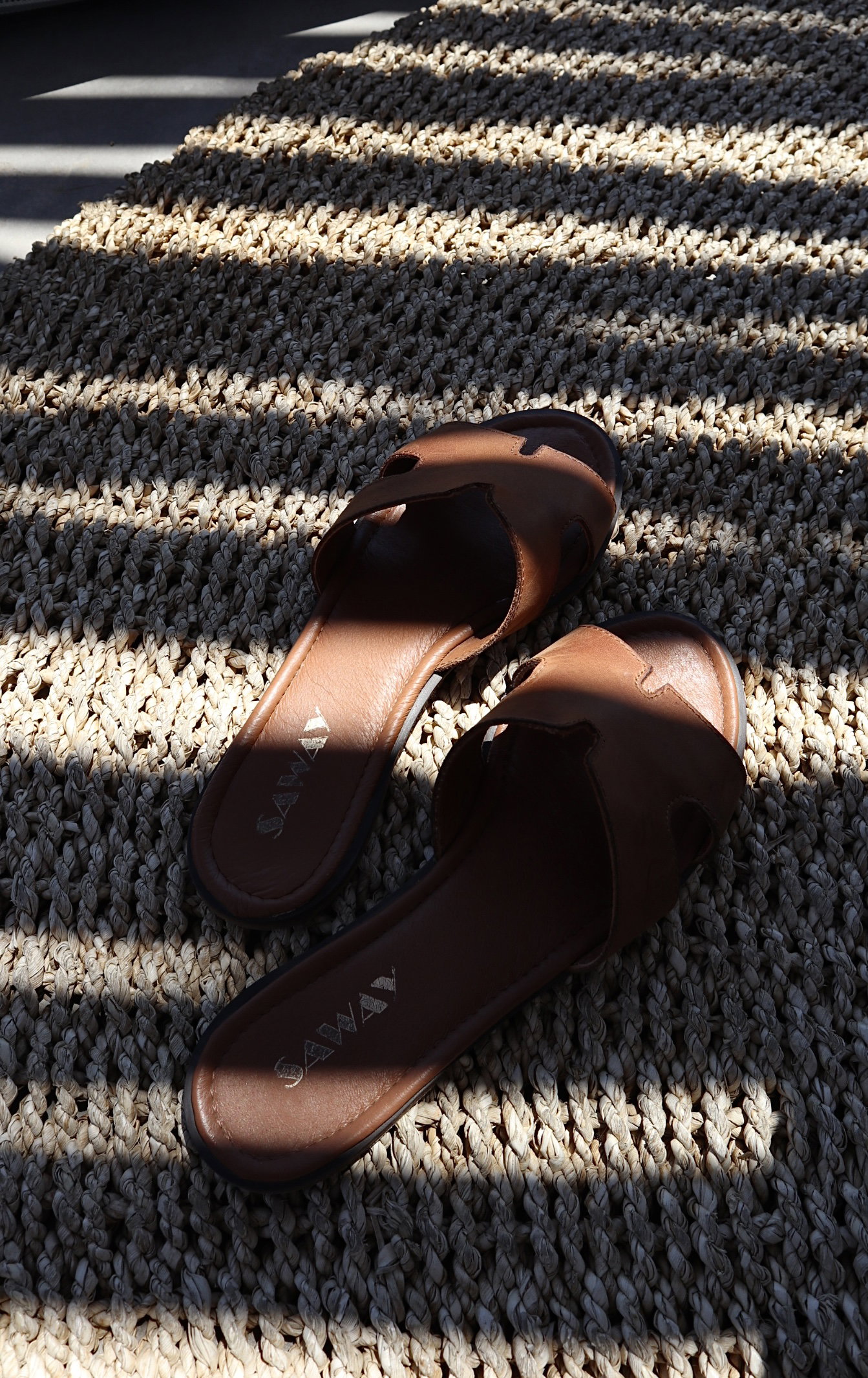 Pair of brown leather slide sandals on a woven beige rug with striped sunlight and shadow pattern