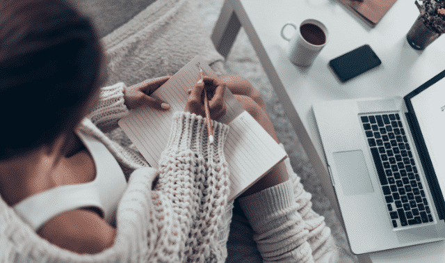 Woman sitting at computer writing her family history in a notebook.