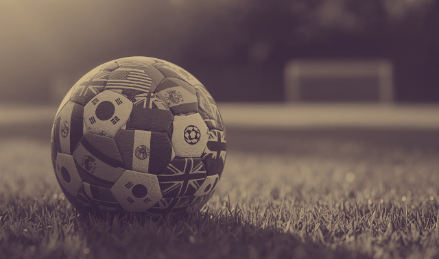A soccer ball decorated with various national flags rests on a grass field with a goalpost visible in the background.