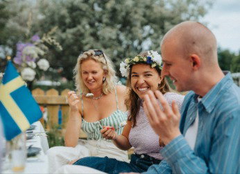 Ein Mann und zwei Frauen sitzen am Tisch und Essen anlässlich des Midsommar-Festes Kuchen. Jetzt Flüge nach Schweden buchen.