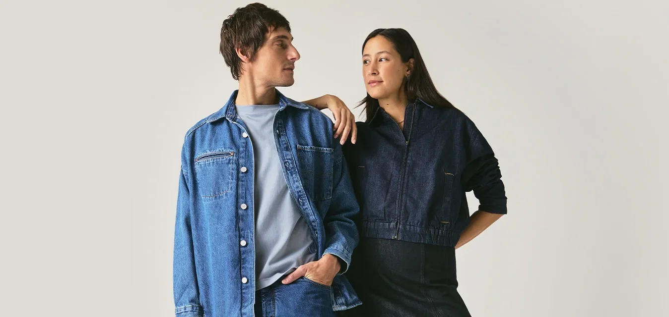 Mann und Frau stehen sich gegenüber, beide tragen dunkle Denim-Outfits mit schwarzen Schuhen vor einem einfarbigen hellgrauen Hintergrund.