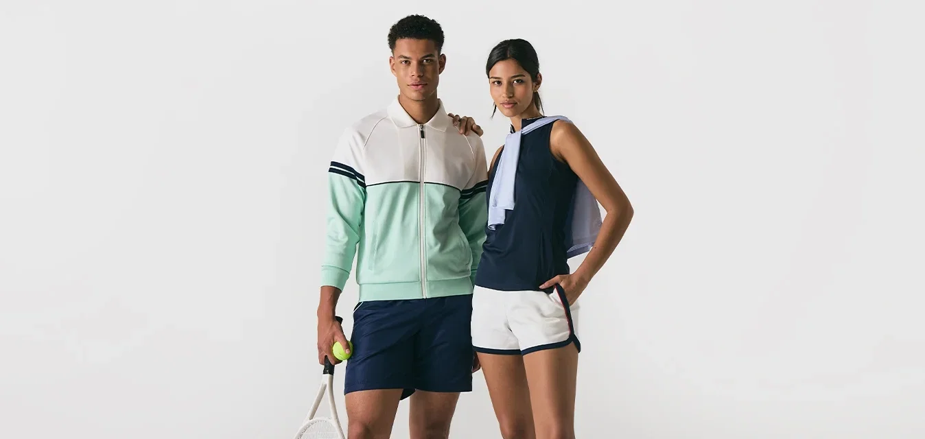 Junger Mann und junge Frau in Tennisbekleidung stehen nebeneinander, der Mann hält einen Schläger und einen Ball, die Frau hat die Hand in der Tasche und trägt eine Jacke über der Schulter.
