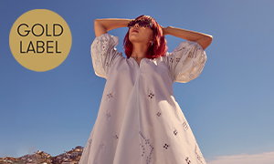 Person wearing a loose, white, embroidered dress and sunglasses stands outdoors under a blue sky. A gold circle in the corner contains the text, “Gold Label”.