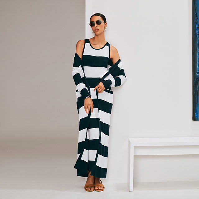 Person wearing a black and white striped dress with sunglasses, leaning against a wall near a white bench.