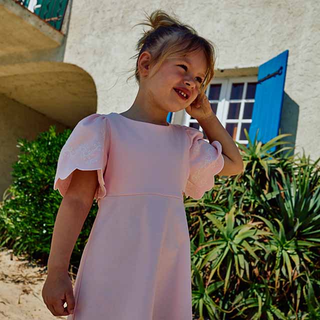 A young girl in a pink dress stands outdoors, smiling, with plants and a building in the background.