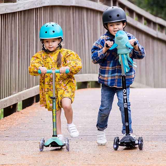 Two children wearing helmets ride scooters on a bridge.