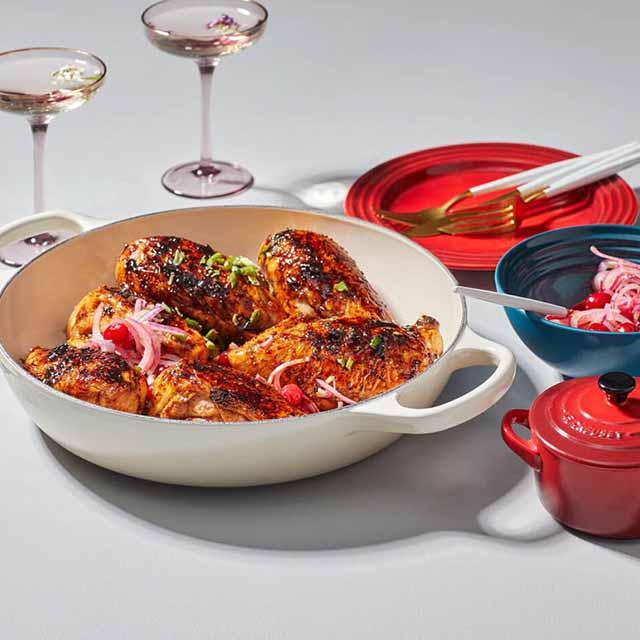 A dish with cooked chicken topped with onions in a white pan, accompanied by plates, cutlery, and two glasses on a table.