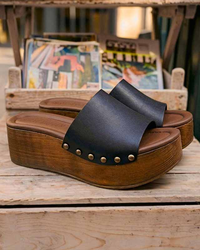Dark blue leather clogs with wooden platform heels on a rustic surface, magazines in the background.
