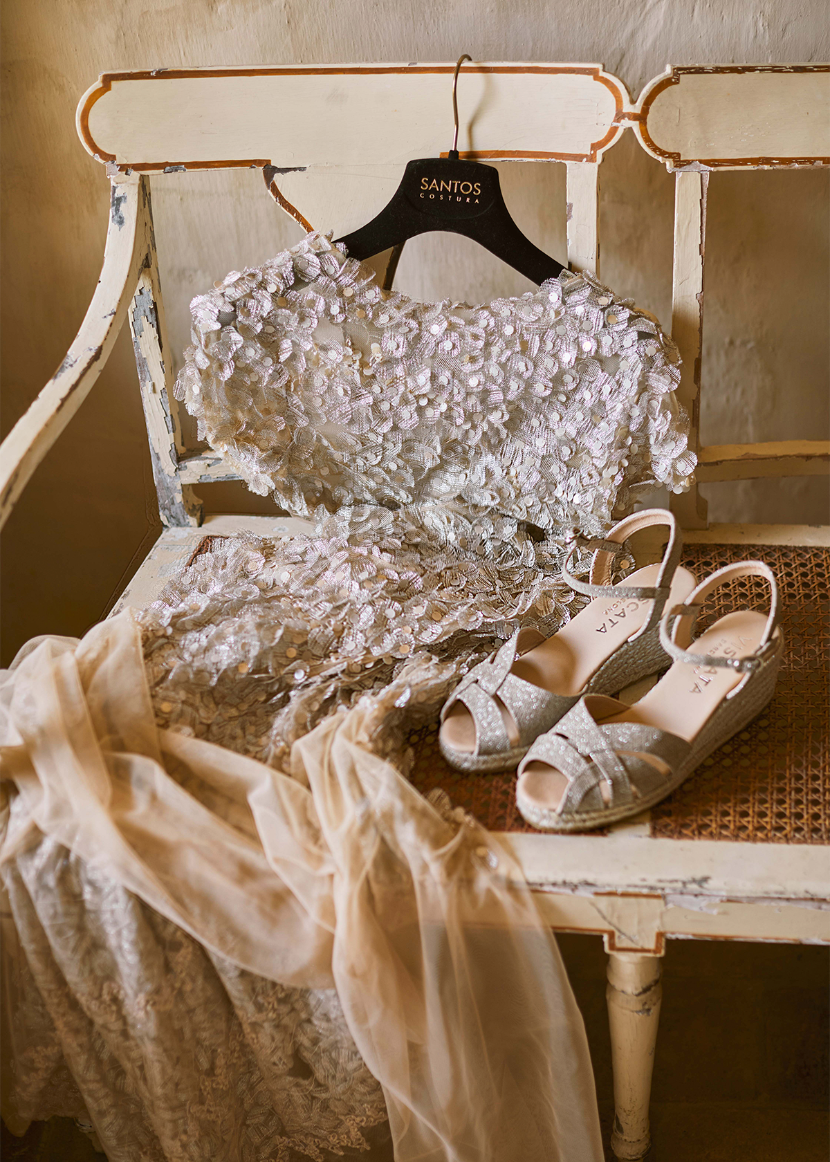 Champagne glitter Laia espadrille wedge sandals displayed beside an embellished occasion dress on a vintage chair in a warm, softly lit interior.