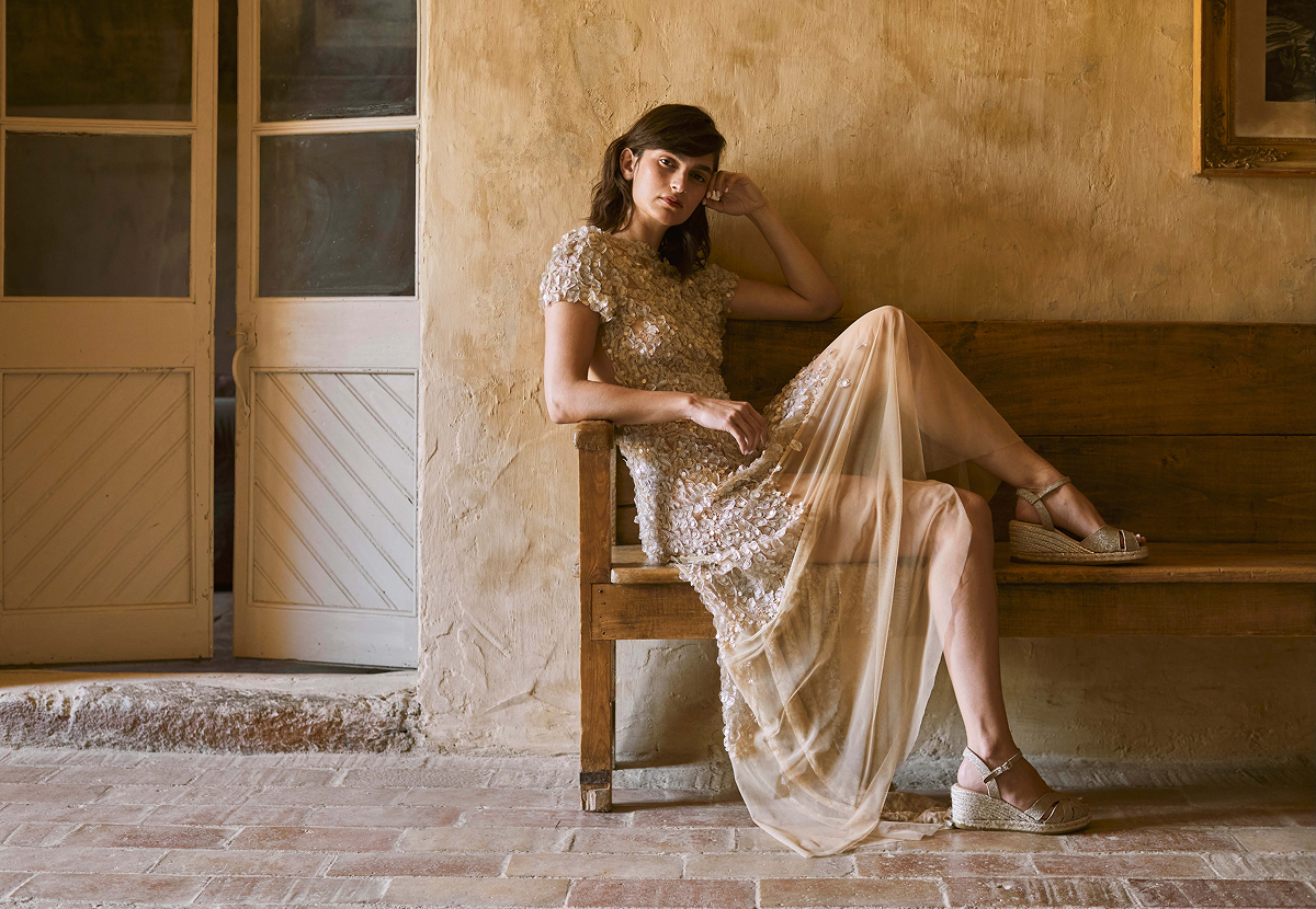 Woman seated on a rustic wooden bench in a soft embellished dress, wearing silver Laia espadrille wedge sandals in a warm Mediterranean interior.