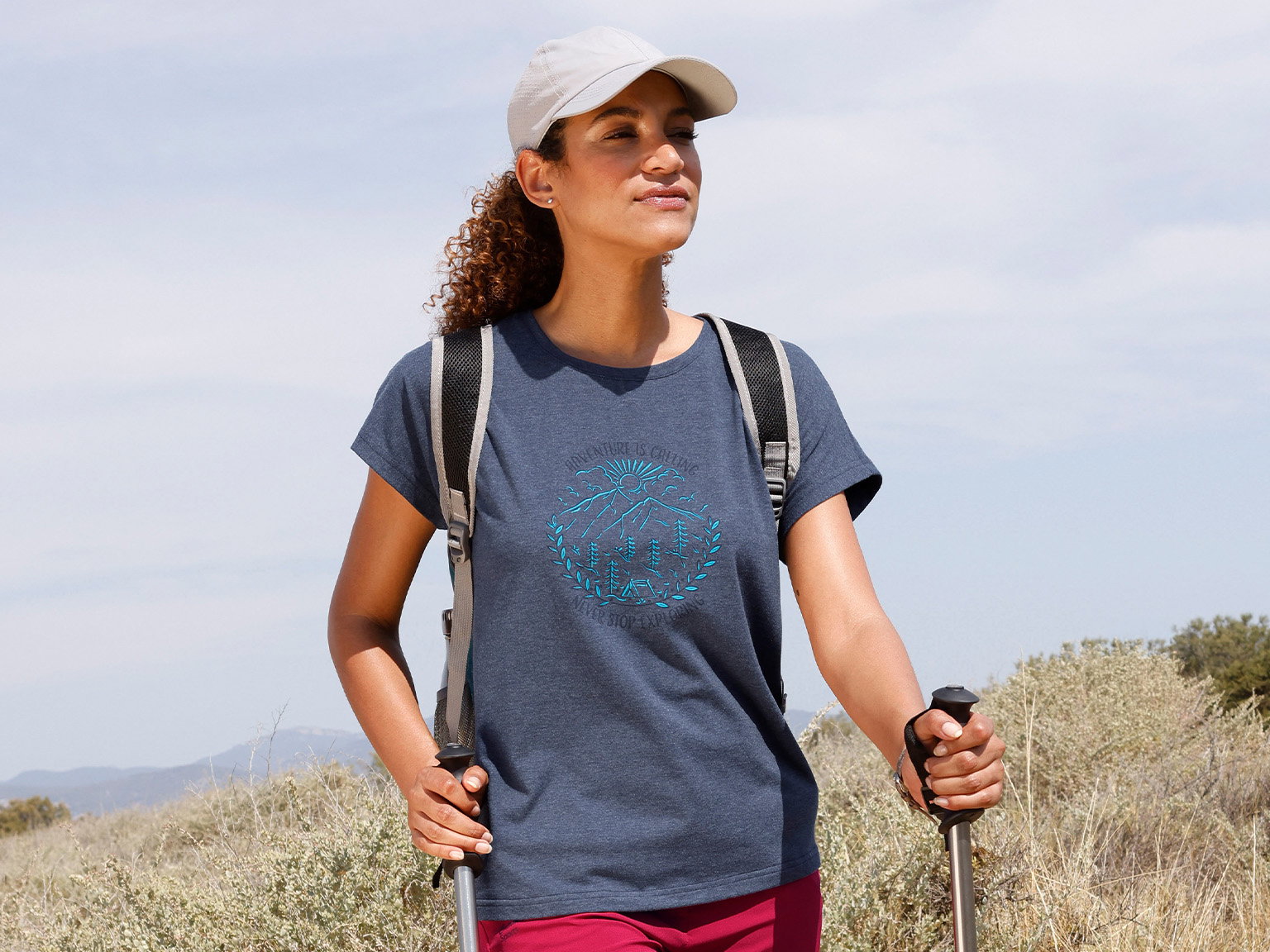Ein Model mit lockigem Haar trägt ein blaues T-Shirt, eine weiße Kappe und einen Rucksack, hält zwei Wanderstöcke und steht in einer offenen Landschaft. 