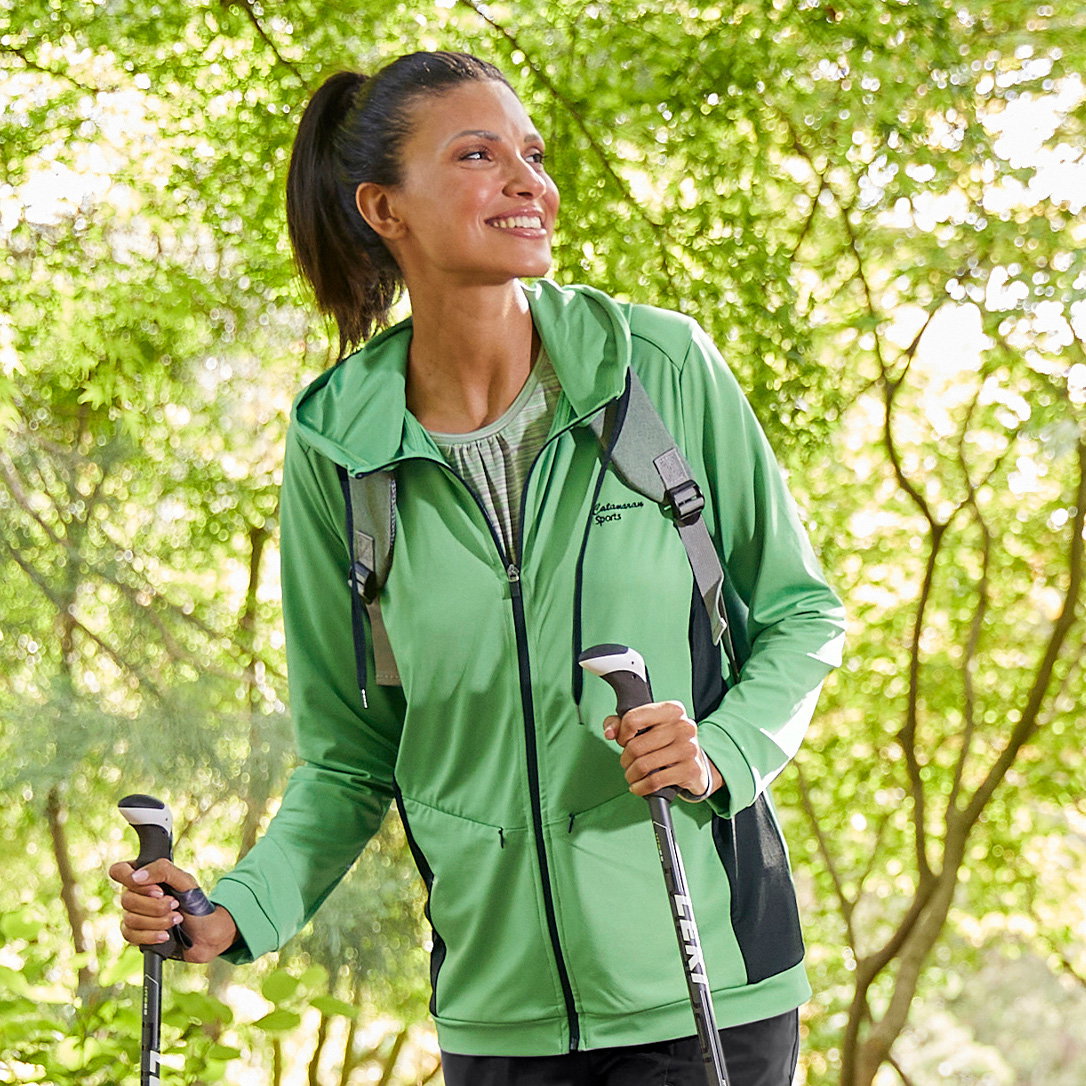 Ein Model mit Pferdeschwanz trägt eine grüne Sportjacke und hält zwei Wanderstöcke, steht im Wald bei Tageslicht.