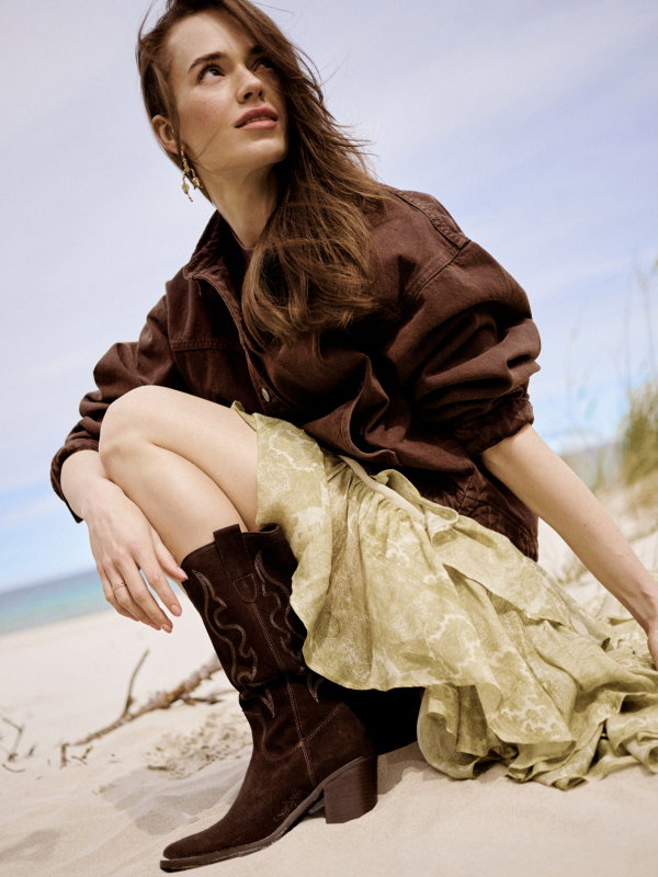 Woman in brown jacket and boots sitting on beach, wearing green skirt.