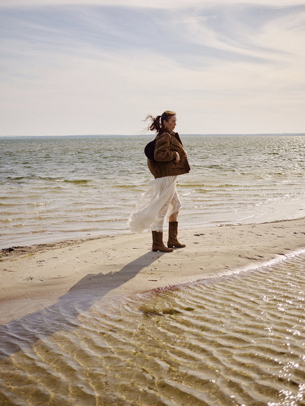 Woman in brown jacket and boots walking along a sandy beach by the sea