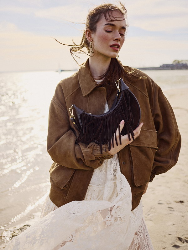 Woman in brown jacket and lace dress holding a fringed bag on a beach at sunset