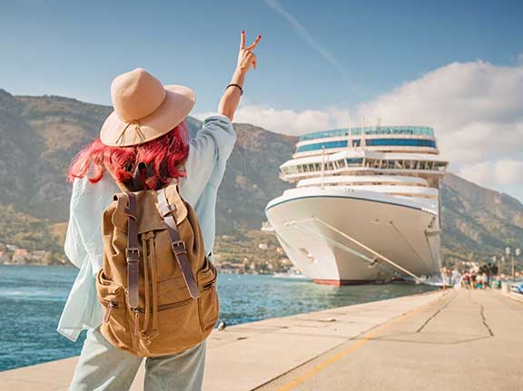 Frau mit roten Haaren, Hut und Rucksack läuft an einem sonnigen Tag auf einem Steg zu einem großen Kreuzfahrtschiff zu.