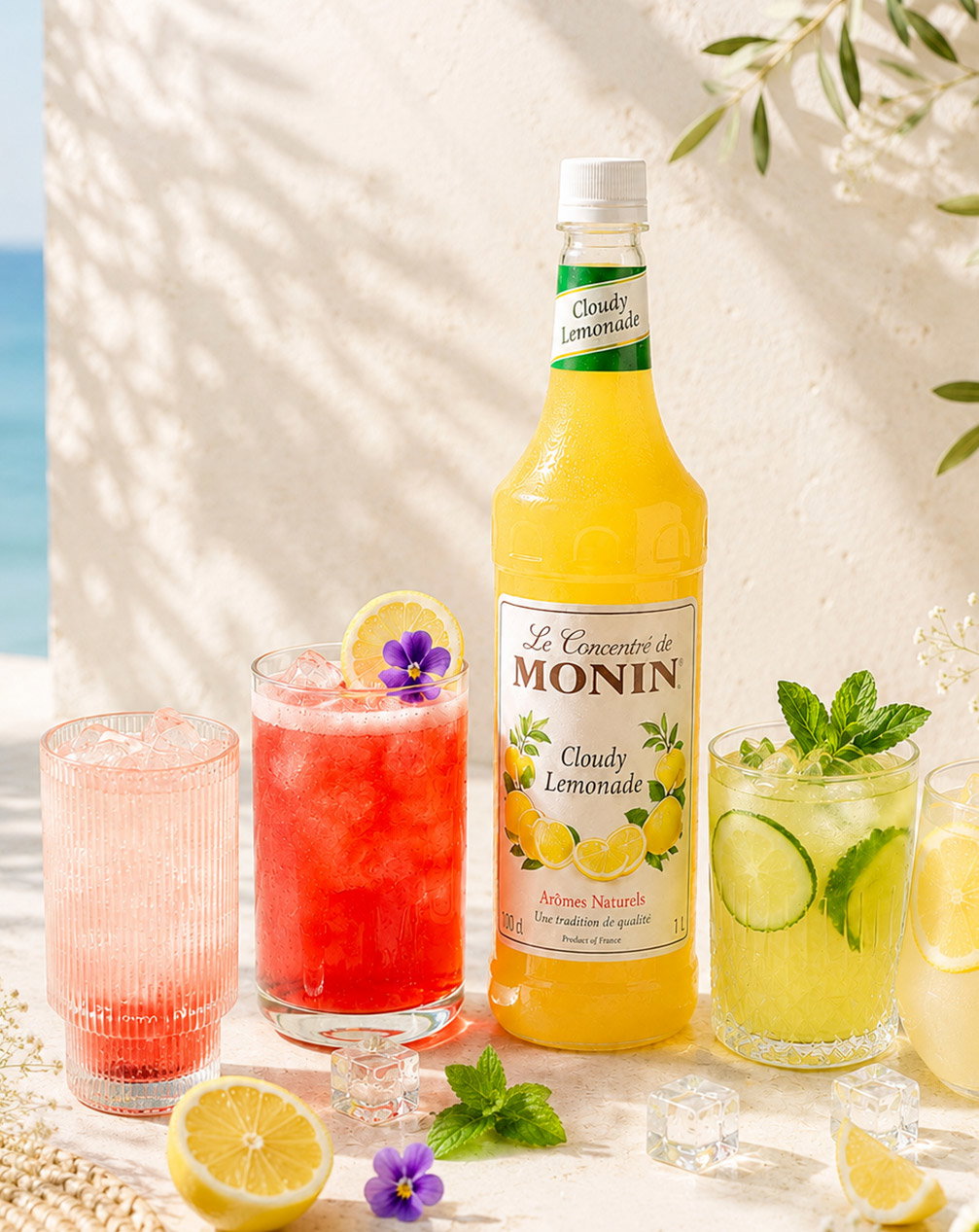 Monin Cloudy Lemonade bottle with three colourful cocktails garnished with lemon, mint and edible flowers by a sunny seaside