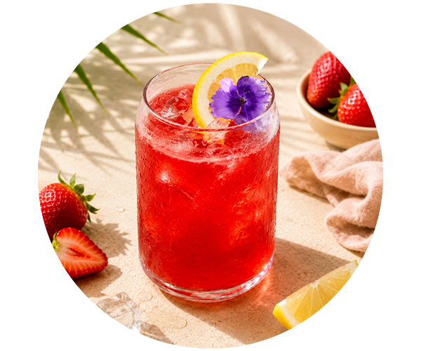 Iced strawberry lemonade in a glass, garnished with a lemon wedge and purple edible flower, fresh strawberries and ice nearby