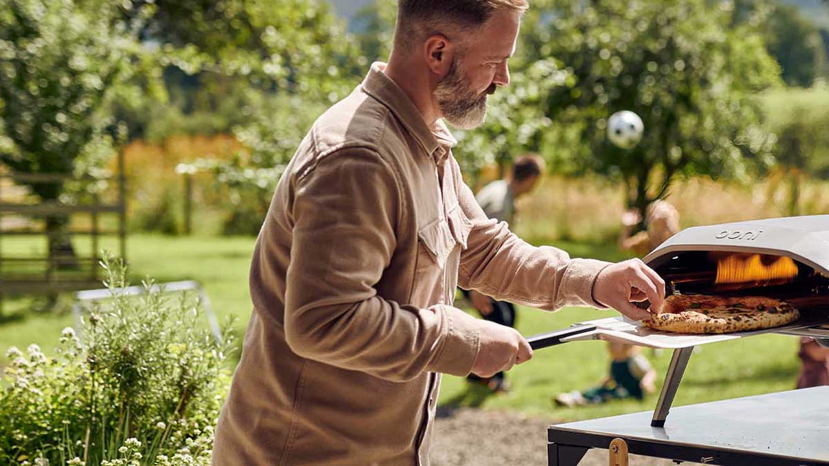 A man cooking a pizza outside in his garden whilst people play soccer in the background. 